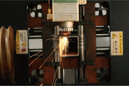 Square steel pipe being processed by vertical laser cutting head, showing intense spark output and clamping system
