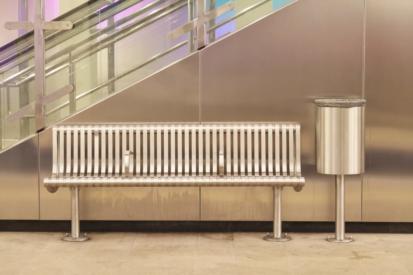 Sleek stainless steel bench next to a matching trash can against a metal-clad wall and glass staircase